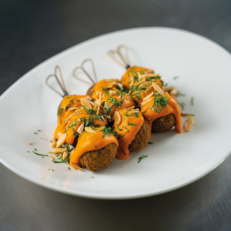 Immagine degli spiedini di polpette vegetali, accompagnati dalla salsa al peperone e decorati con prezzemolo tritato e mandorle tostate, serviti su un piatto elegante